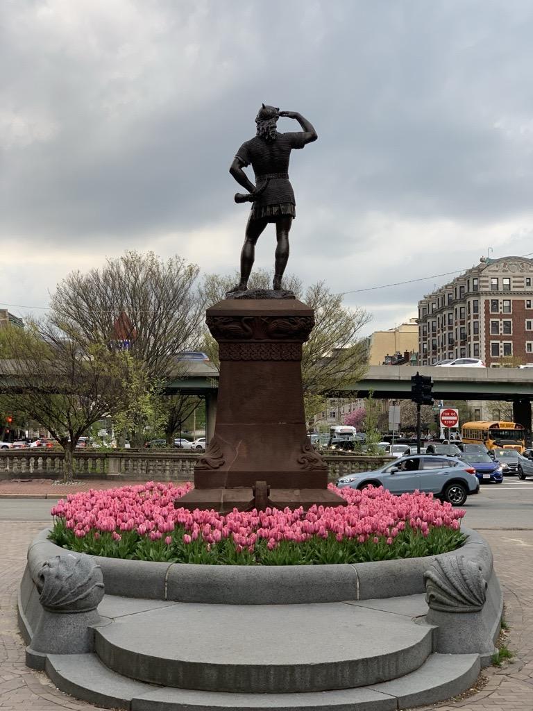 Photograph of bronze statue depicting a man on a stone plinth, taken from the backside view, surrounded by pink tulips. Behind the statue is an overpass with cars